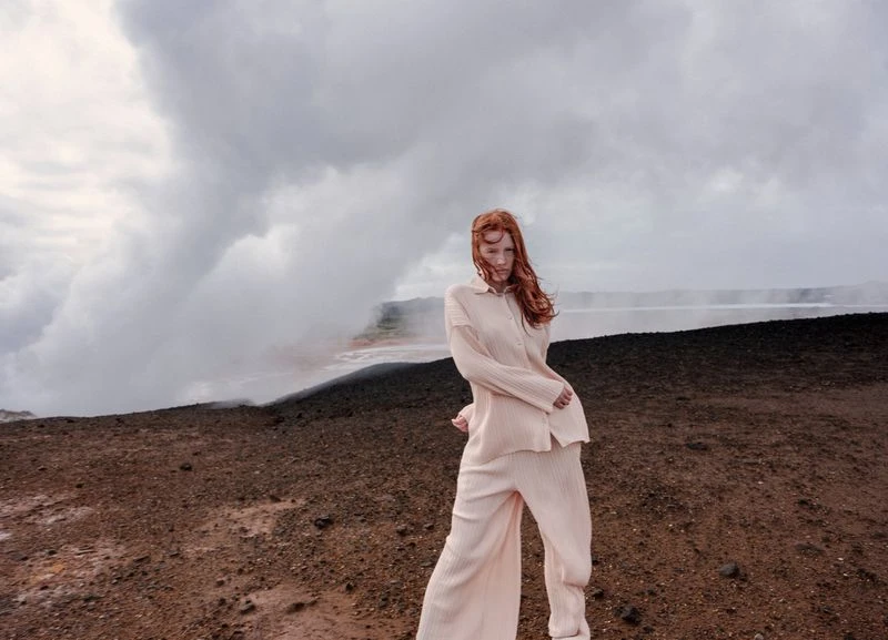 Fashion photography showcasing a young woman with red hair in a beige outfit, posing against a volcanic landscape with steam in background.