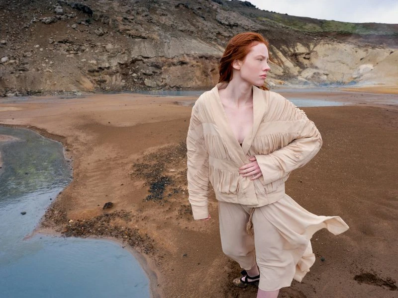 Fashion shot of a redhead woman in a jacket and skirt posing in a volcanic landscape with muted beige and blue tones.
