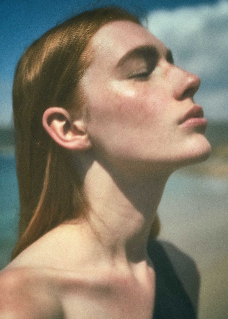 Close-up portrait of a red-haired woman with closed eyes, bathed in sunlight on a beach with a clear blue sky and water backdrop.