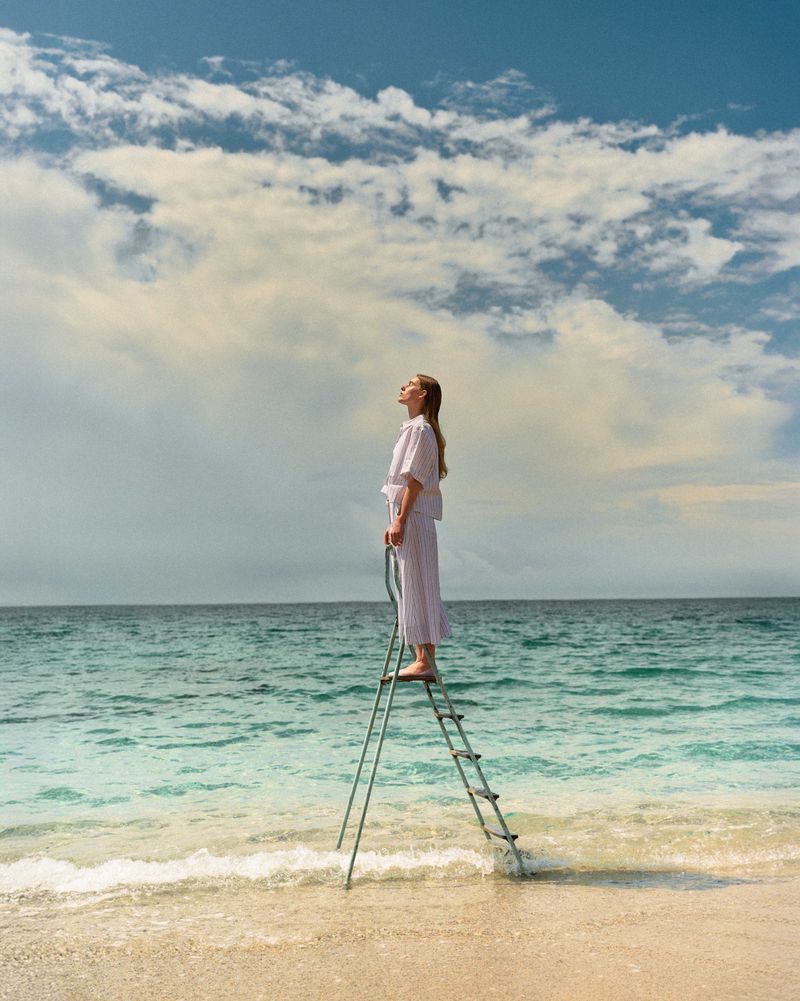 Woman in white outfit stands on ladder in ocean on sandy beach with cloudy blue sky overhead in a medium, eye-level shot.