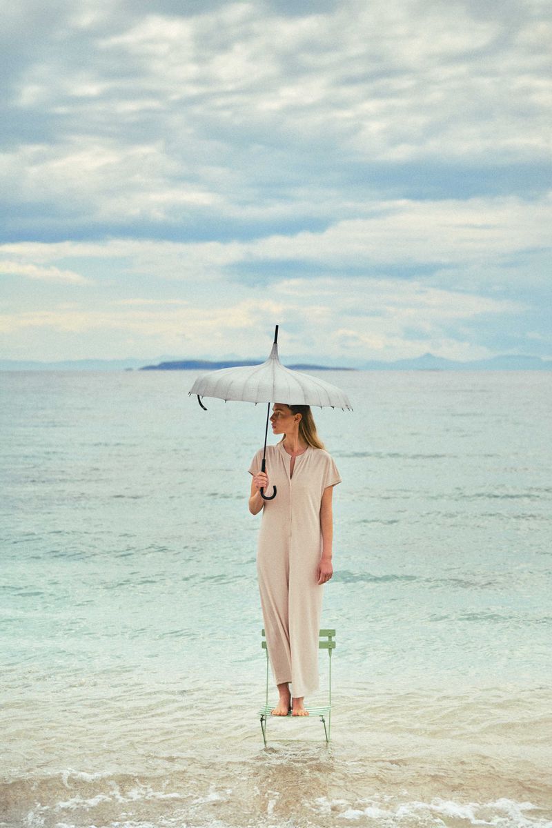 Adult woman stands on a chair in the sea holding a parasol. Sky is cloudy and an island is visible on the horizon in the distance.