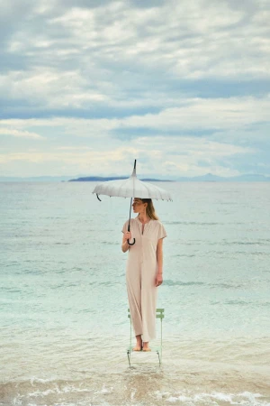 Adult woman stands on a chair in the sea holding a parasol. Sky is cloudy and an island is visible on the horizon in the distance.