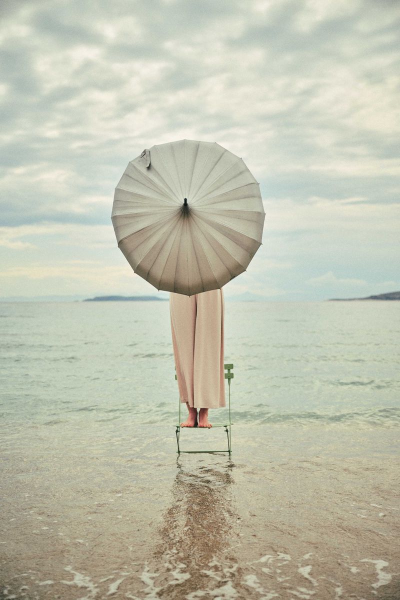 A person stands on a green chair in the ocean, holding a light grey umbrella that covers their head and torso against a cloudy sky.