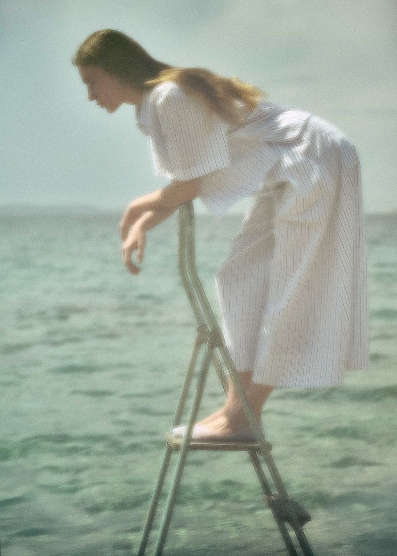 A young woman in a long white dress stands on a silver metal ladder in the shallow water, with a blurry sky and sea background.