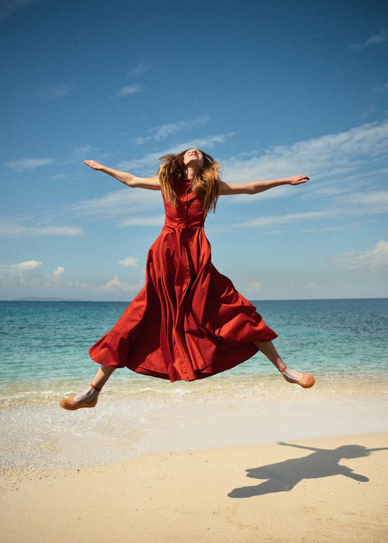 A woman in a red dress jumps barefoot on a sandy beach with her arms outstretched against a sunny blue sky and ocean backdrop.