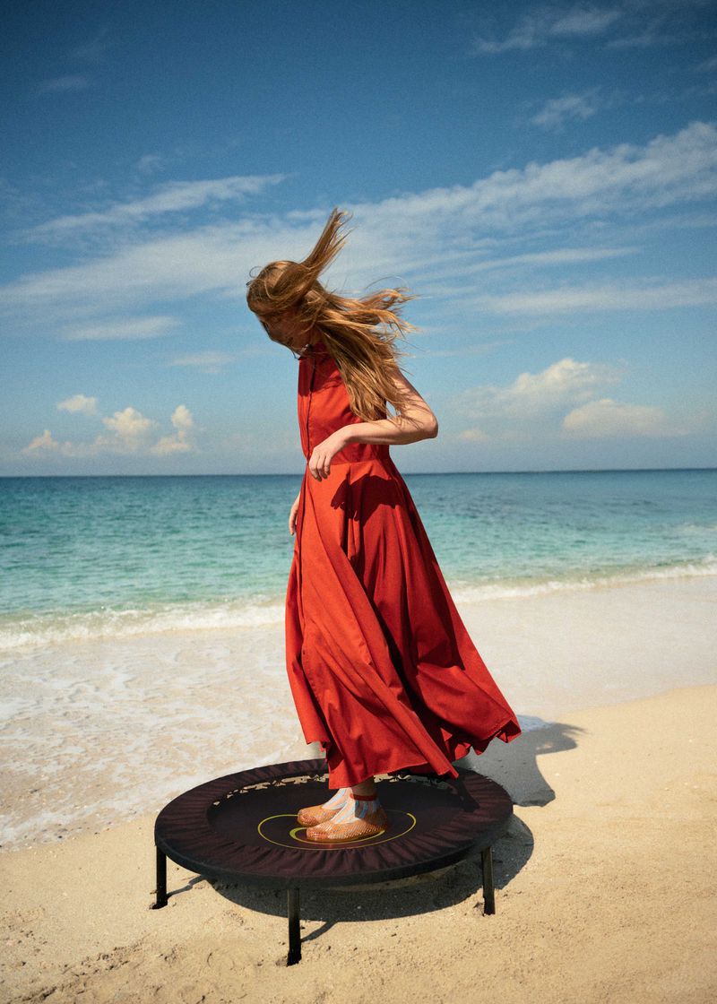 Adult woman with long red hair in a red dress is standing on a trampoline at the beach in front of the ocean and blue sky.