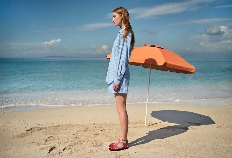 Woman in a blue striped outfit poses in profile on a sunny beach near an orange parasol with blue ocean and sky in the background.