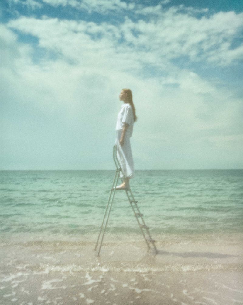 Woman stands on a ladder at the beach, wearing white clothing, looking up at the clouds in the sky with the sea in the background.