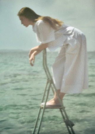 Woman in a white striped short-sleeve dress leaning forward on a ladder in calm coastal sea water.