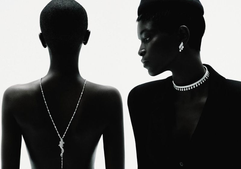 Two Black women in a studio setting wearing formal black tops and diamond jewelry necklaces and earrings against a bright white background.
