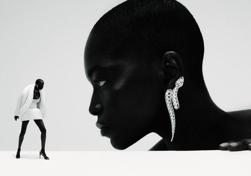 Black and white fashion shot of a Black woman's profile wearing a diamond serpent earring next to a miniature figure in a white mini dress.