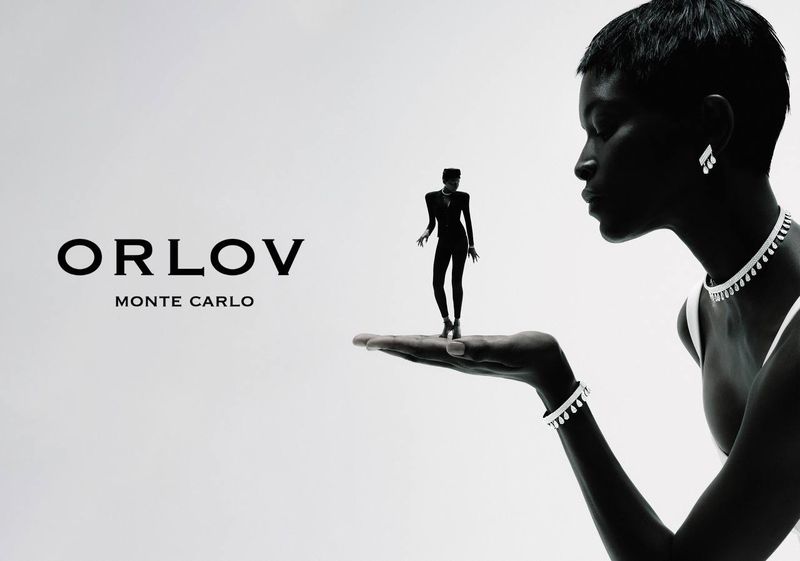 Black and white portrait shot of an adult female wearing jewelry, miniature woman on her hand, and Orlov Monte Carlo logo.