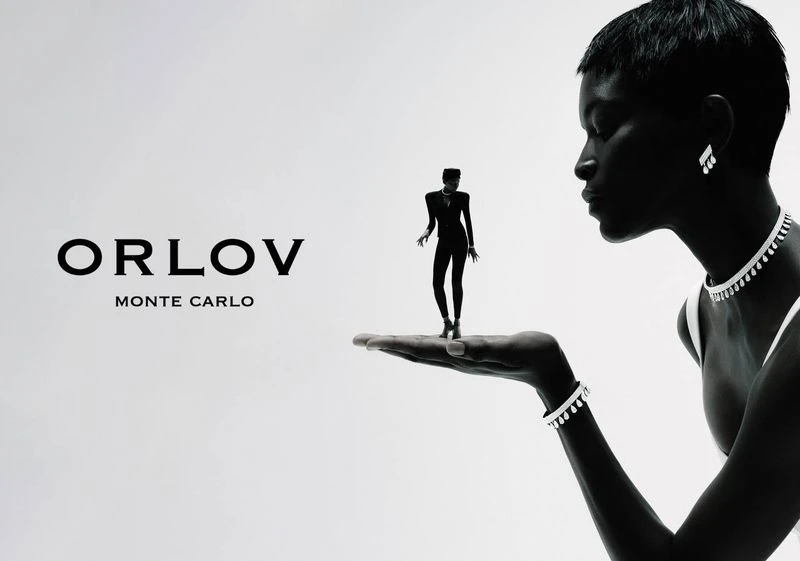 Black and white portrait shot of an adult female wearing jewelry, miniature woman on her hand, and Orlov Monte Carlo logo.