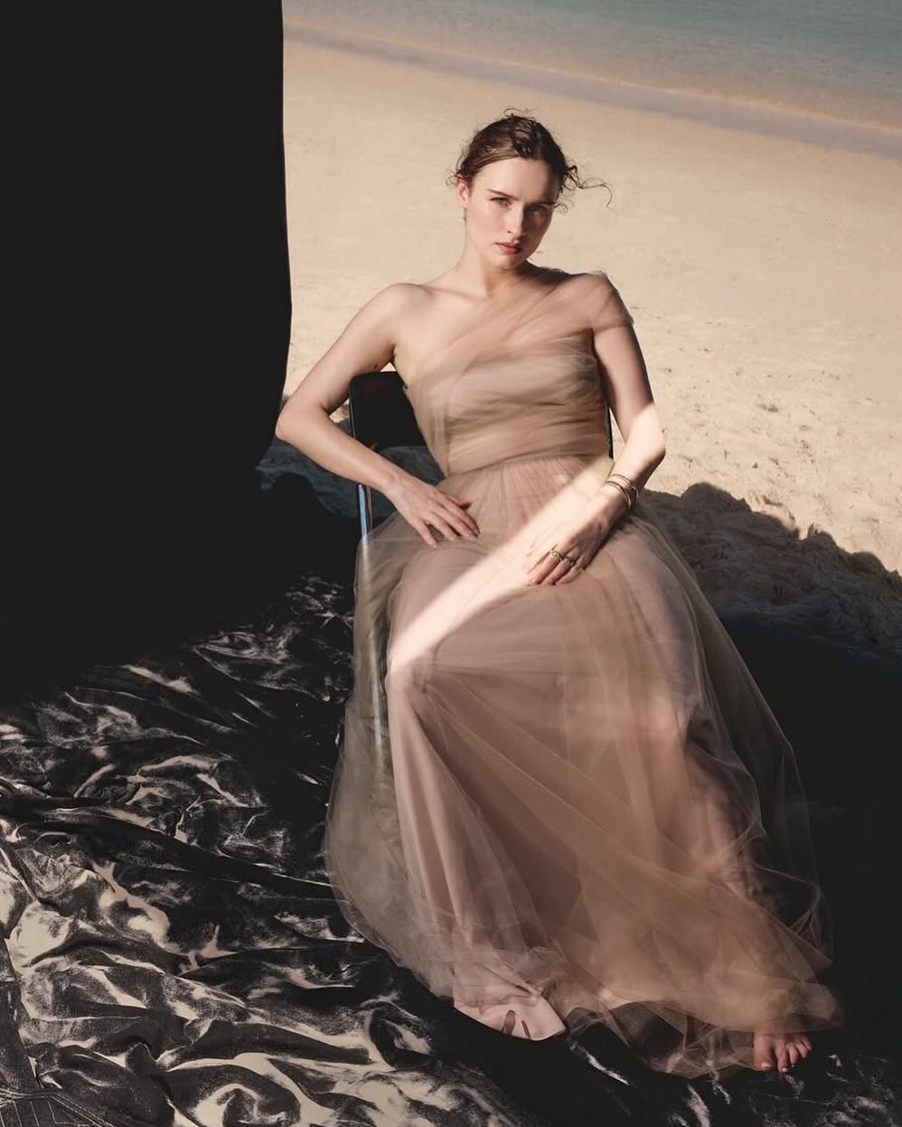 Woman in an elegant nude dress poses seated on a beach, a play of light and shadow on the ocean shore in the background.