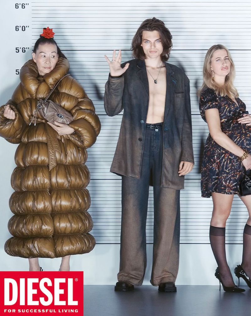 Three models stand in a lineup against a height chart in a studio. The woman on the left wears a large puffer coat, the man is shirtless, and the woman on the right wears a dress.