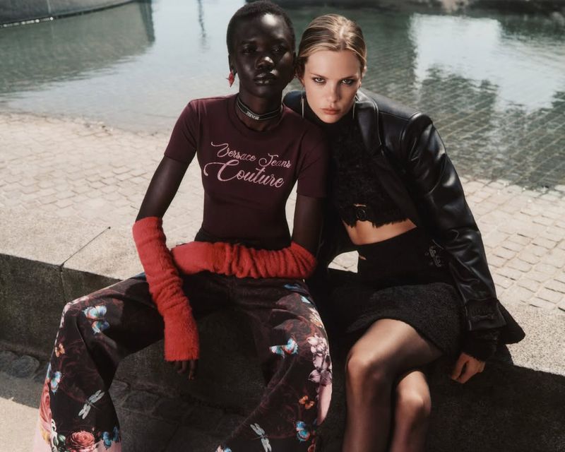 Two fashion models pose on a stone ledge in urban setting. One woman wears a maroon t-shirt and floral pants, the other wears black leather jacket.