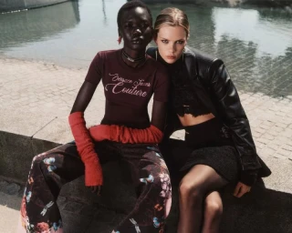 Two fashion models seated outdoors by a stone pool ledge wearing a burgundy graphic tee, floral pants, and a black leather jacket.