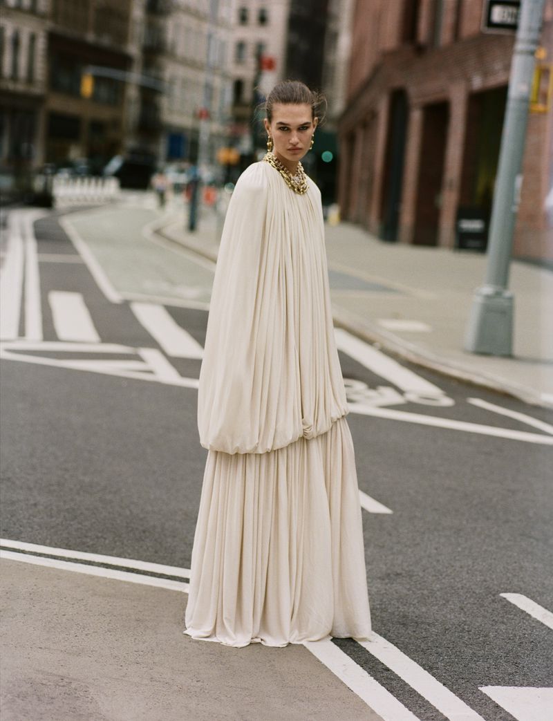 Woman in flowing white gown and gold jewelry stands on a city street crosswalk with buildings in the background on an overcast day.