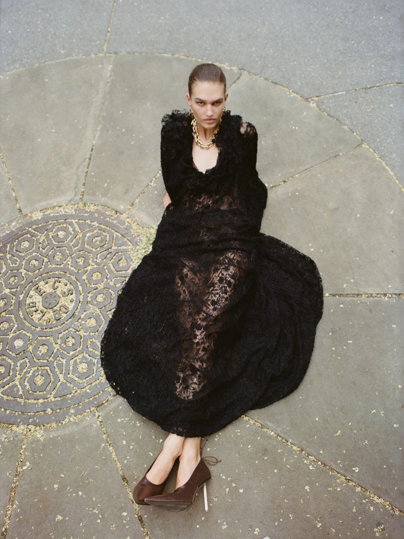Overhead fashion shot of a woman in a black lace dress and brown heels sitting on a circular stone surface with decorative patterns.
