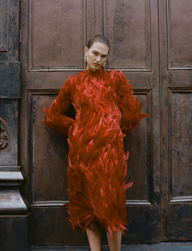 An adult woman poses in a red feathered midi dress in front of a dark brown paneled door on a cloudy day in an outdoor setting.