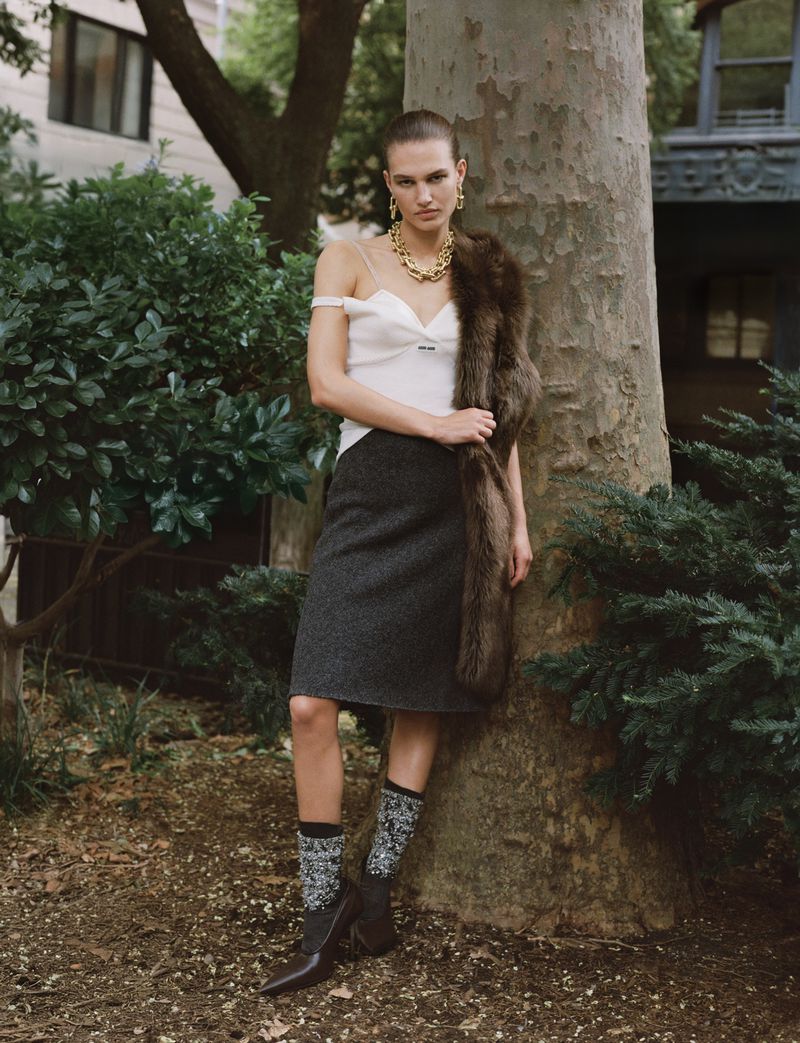 Elegant woman poses outdoors near tree. She wears a white top, gray skirt, jewelry, fur stole, patterned socks, and brown heeled shoes.