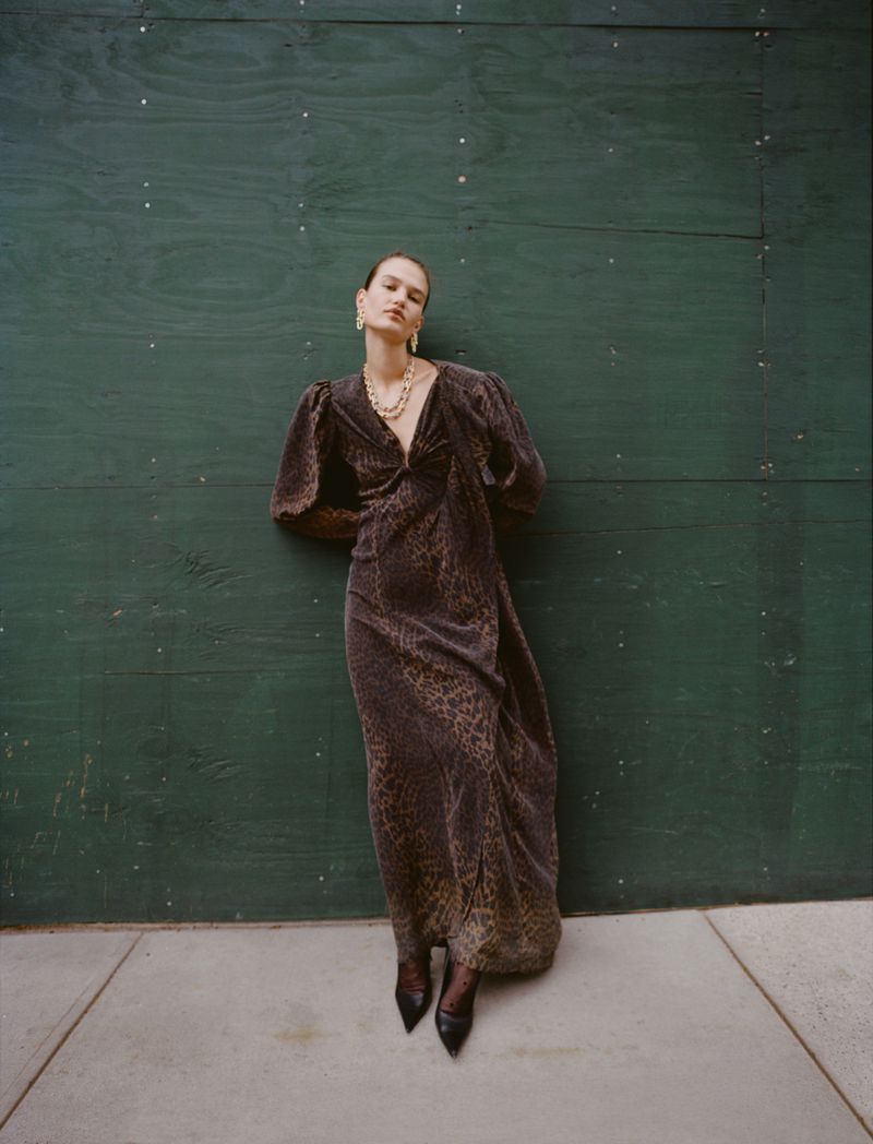 Model Emmi Freeman in a dark patterned maxi dress and black pointed heels leaning against a green wooden wall.