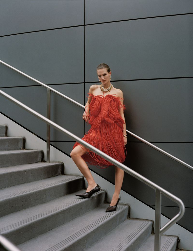 A fashionable adult woman is walking up gray stairs in an orange fringed dress and black heels, wearing a necklace and earrings.