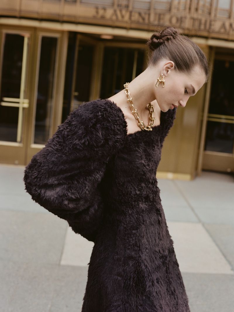 Portrait of a woman in a black textured dress and gold jewelry stands outdoors in front of an urban building with golden doors.