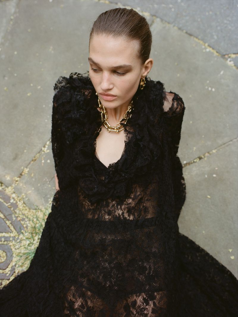 High-angle shot of a female fashion model in a long black lace dress and gold chain necklace, set against a stone background.