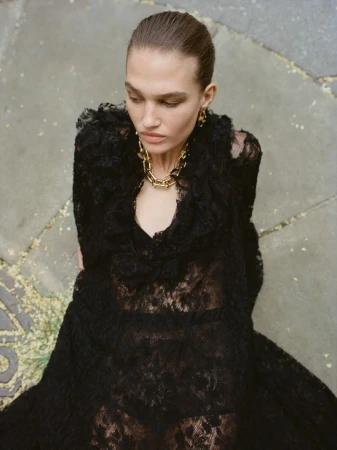 High-angle shot of a female fashion model in a long black lace dress and gold chain necklace, set against a stone background.