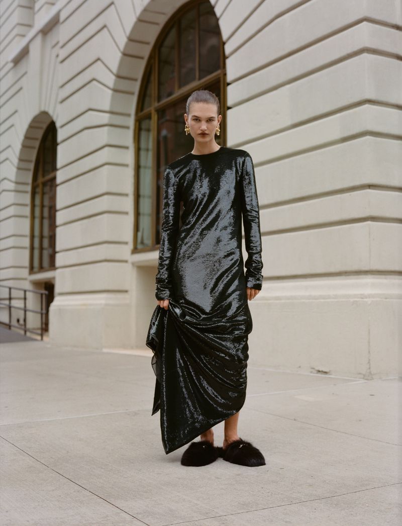 A woman in a black dress poses outdoors against a white building with arched windows and gray pavement on a cloudy day.