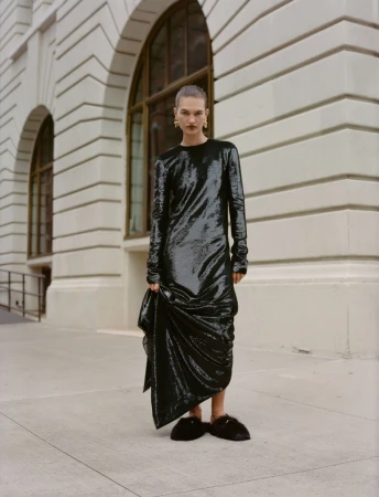 A woman in a black dress poses outdoors against a white building with arched windows and gray pavement on a cloudy day.