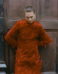 Model Emmi Freeman posing in a vibrant red feathered long-sleeve dress against a weathered brown paneled door.