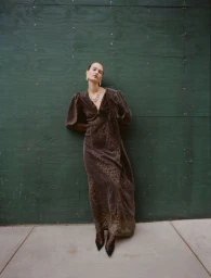 Model Emmi Freeman in a dark patterned maxi dress and black pointed heels leaning against a green wooden wall.