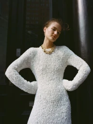 Model wearing a textured white dress and thick gold chain necklace posing against a dark building exterior.