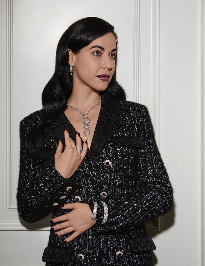 Model Anna Alimani posing in a black tweed blazer and statement diamond jewelry against a white paneled wall.