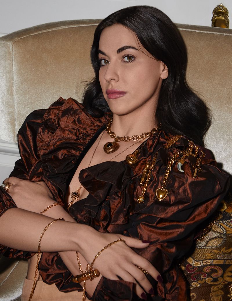Woman in a brown blouse with puffy sleeves, wearing gold jewelry including a necklace and bracelets, sitting on an ornate chair.