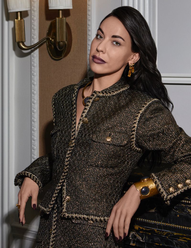 Brunette woman in a tweed jacket and skirt with gold jewellery leaning on a decorative fireplace. Elegant, fashionable attire and posing.