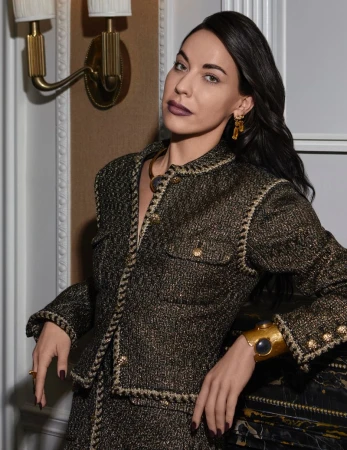Brunette woman in a tweed jacket and skirt with gold jewellery leaning on a decorative fireplace. Elegant, fashionable attire and posing.