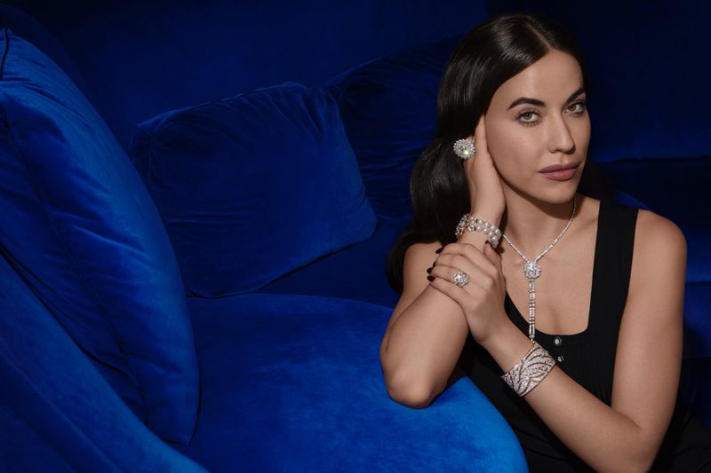 Brunette woman poses on blue velvet couch wearing diamond earrings, necklace, bracelets, and rings in a studio setting.