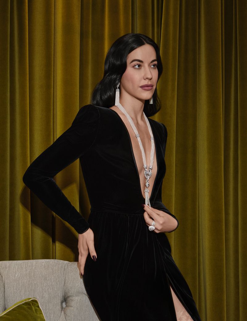 Elegant adult woman with long dark hair wearing a black velvet dress with a slit, statement jewelry, and a full face of makeup.