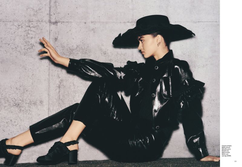 Fashion editorial shot of a stylish woman in an all-black outfit posing against a concrete wall in a studio setting.