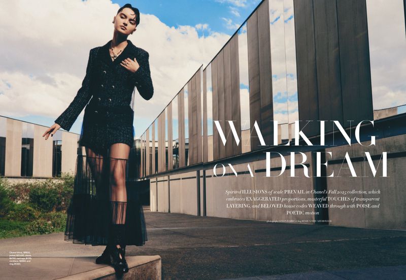 Fashion shot of a model in a dark tweed suit with sheer skirt, posing outdoors against a modern building under a cloudy sky.