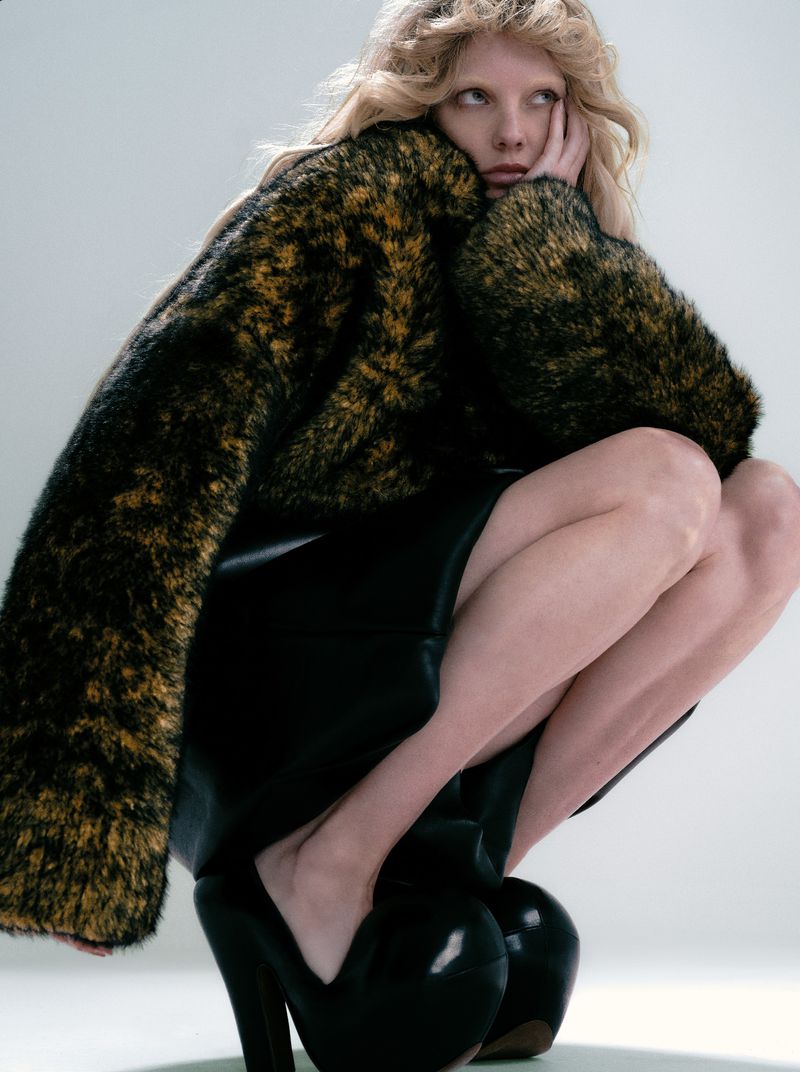 A blonde woman in crouched position wearing a patterned fur coat, black skirt, and high heels in a fashion studio shot.