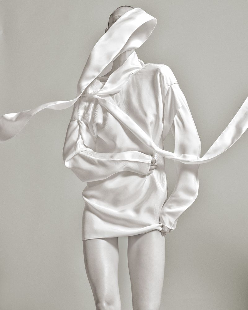 A woman in a draped white outfit poses against a white background, fashion photography. The draped fabric obscures her face.