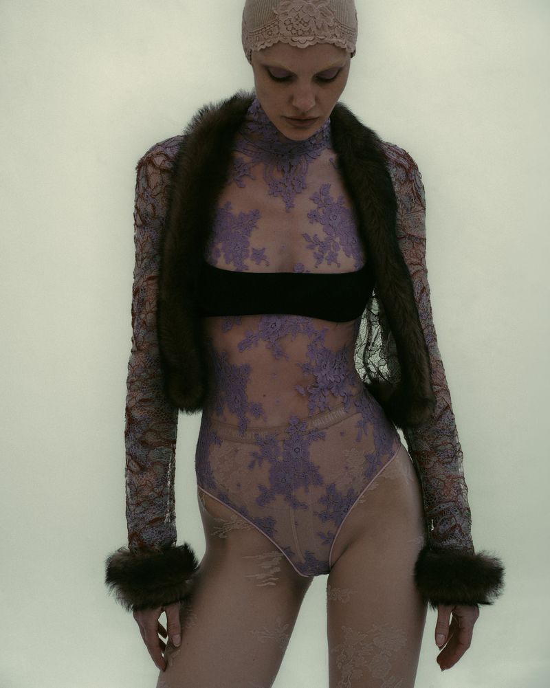 Woman poses in nude lace bodysuit, black bandeau, and fur shawl against a pale backdrop for a fashion editorial shot.