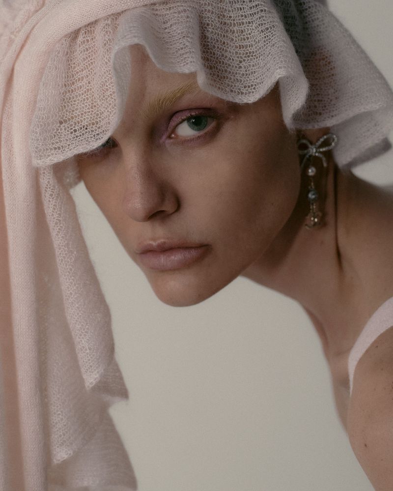 Close-up fashion portrait of a model with light skin and blonde hair wearing a pink hat, bow earrings, and a tank top against light background.
