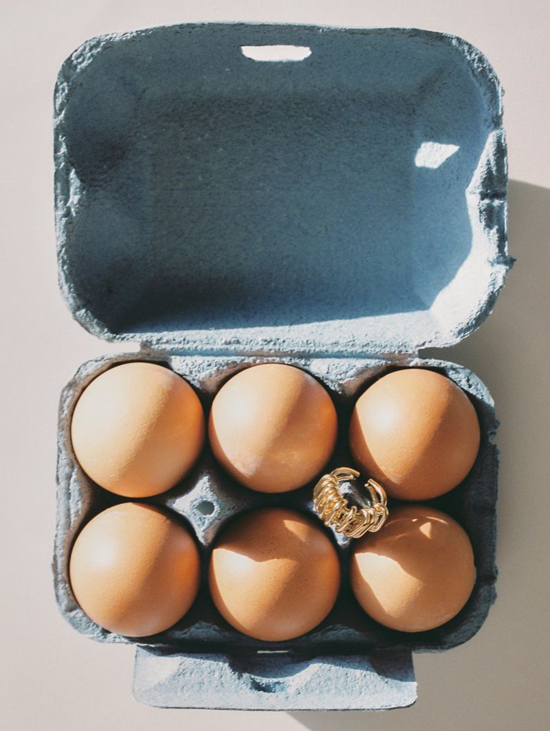 Overhead shot of six brown eggs in a blue egg carton with a gold ring placed on one of the eggs, studio lighting.
