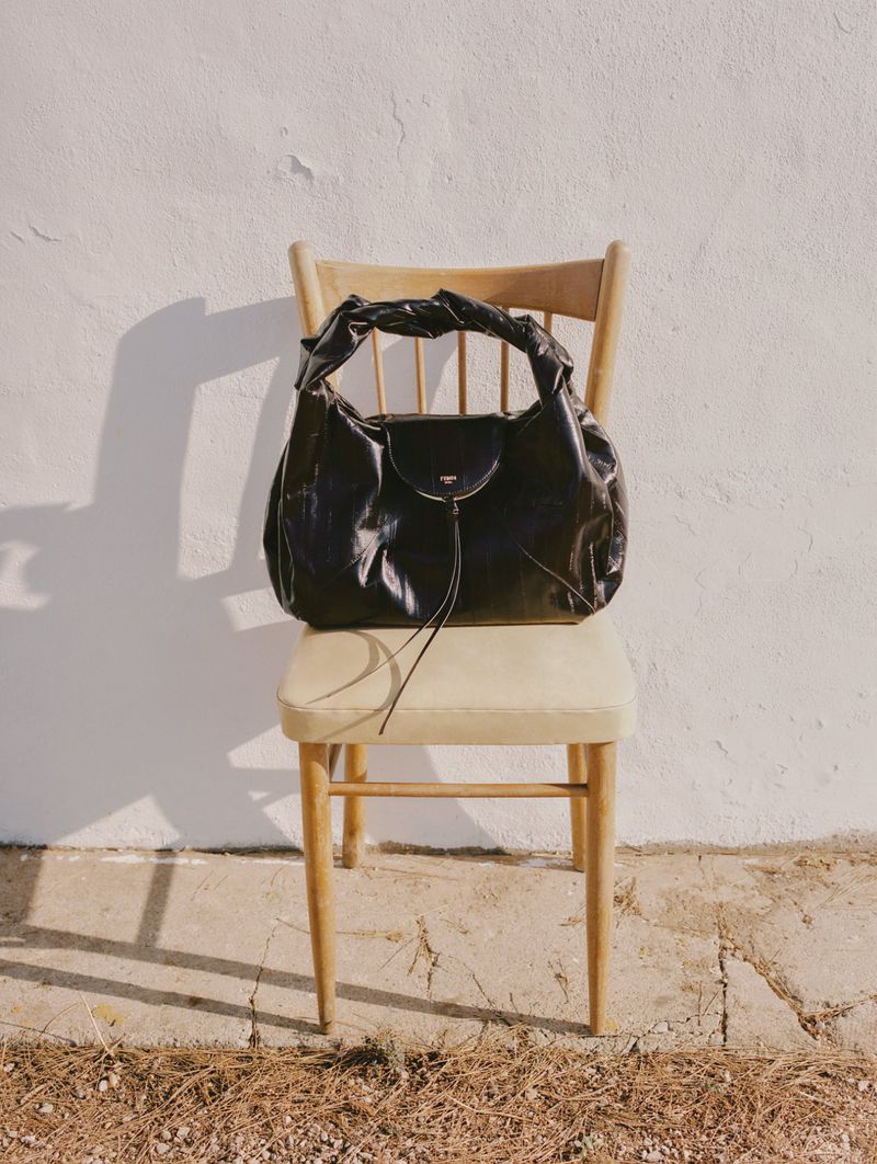 Black leather hobo bag on a vintage wooden chair against a white wall in a sunlit outdoor setting with a concrete floor.