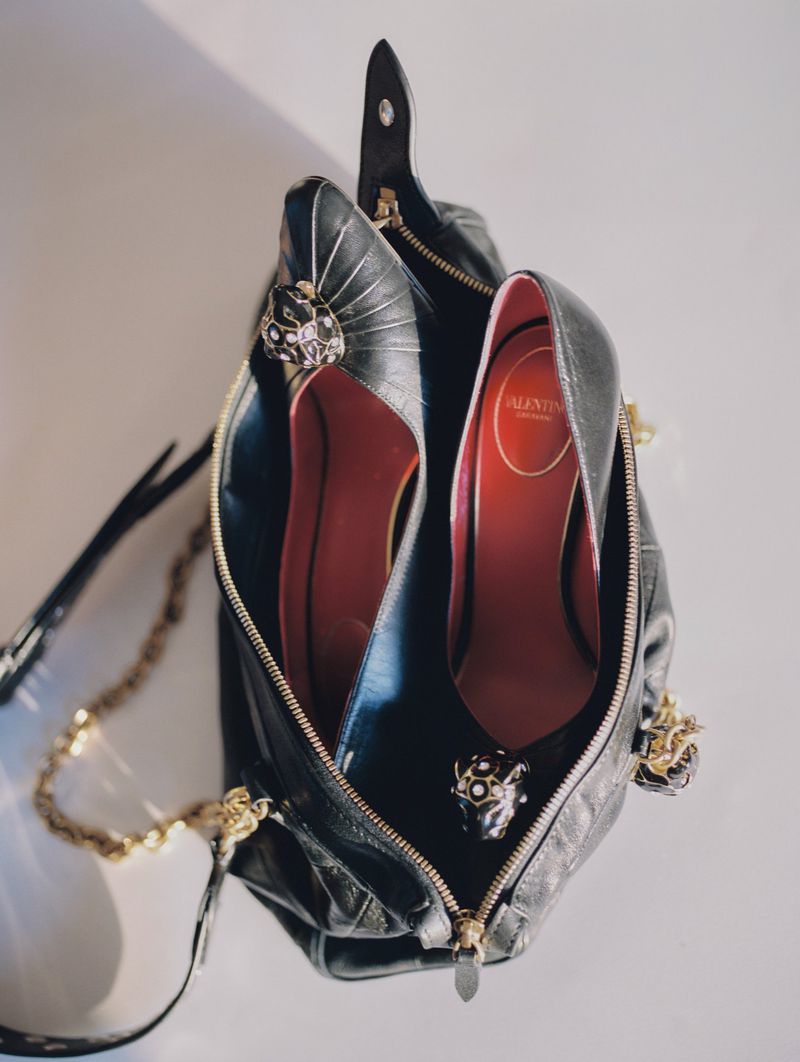 Black leather handbag with elegant black high-heel shoes inside, adorned with decorative skulls, and a gold chain strap on a light surface.